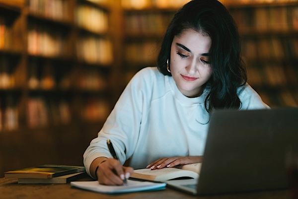 Woman in library, working on a laptop and writing in a notebook